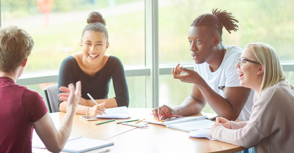 Four students talking, sat around a desk, in the Faculty of Health, Social Care and Medicine.