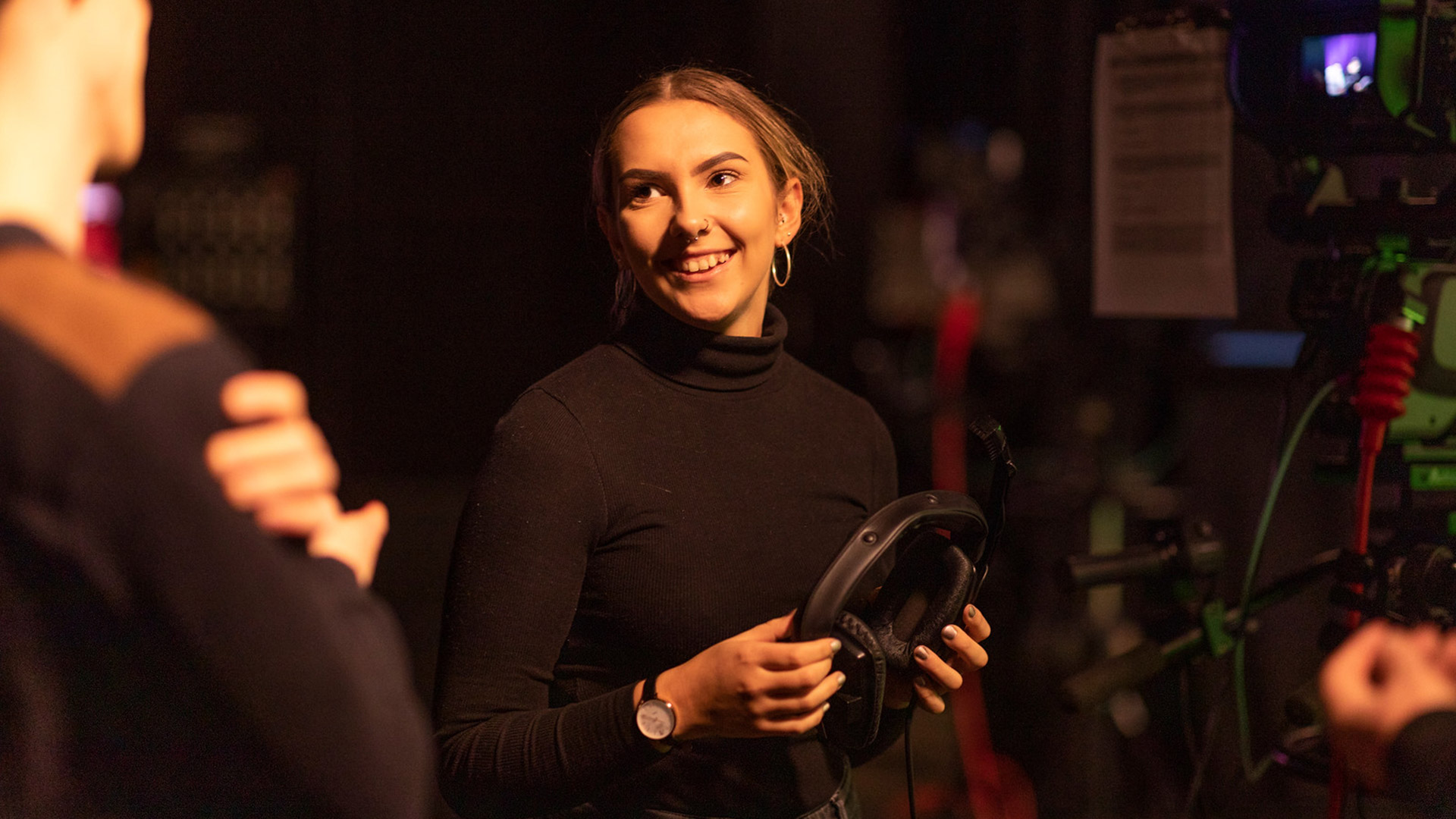 A student smiles as they hold headphones backstage in the TV recording studio