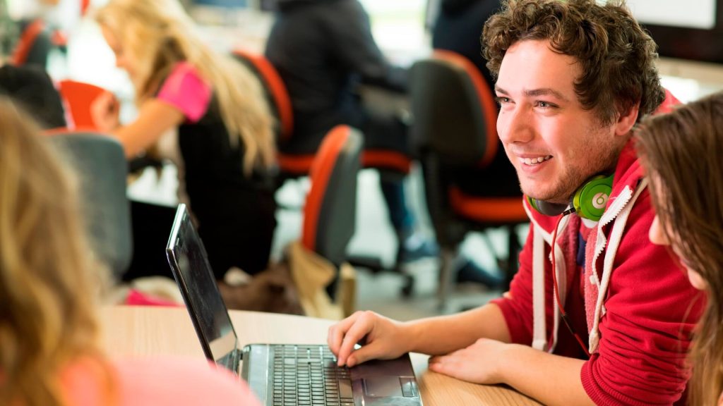 A student, with headphones round his neck, chats with peers while using a laptop.