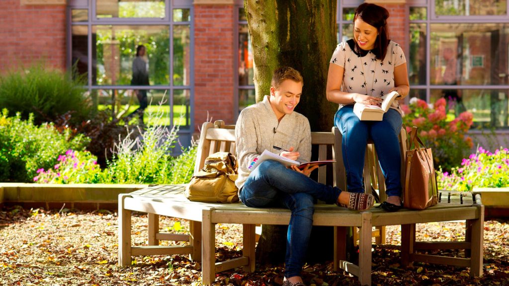 Two students sat on a bench, one reading and the other making notes, in a courtyard by the Main Building.