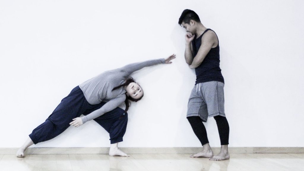 A student leans to one side and stretches their arm forward while another student looks on while learning against a wall.