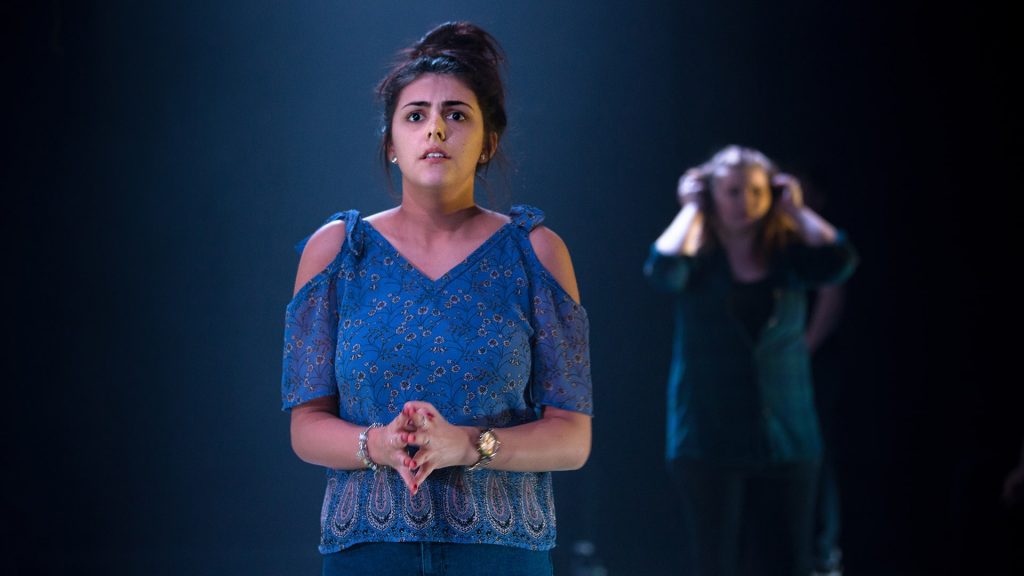 A student participates in a drama performance while one of their peers watches on from behind.