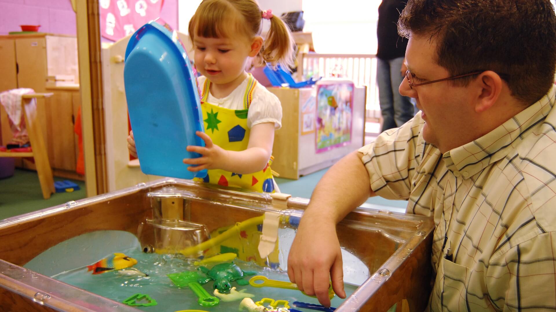 A student plays with a young child as they tip water from a toy boat into a sink full of water and floating toys.
