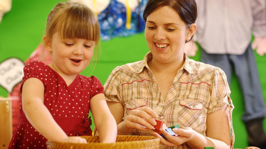 An early years professional plays with a child who is taking small building blocks from a basket.