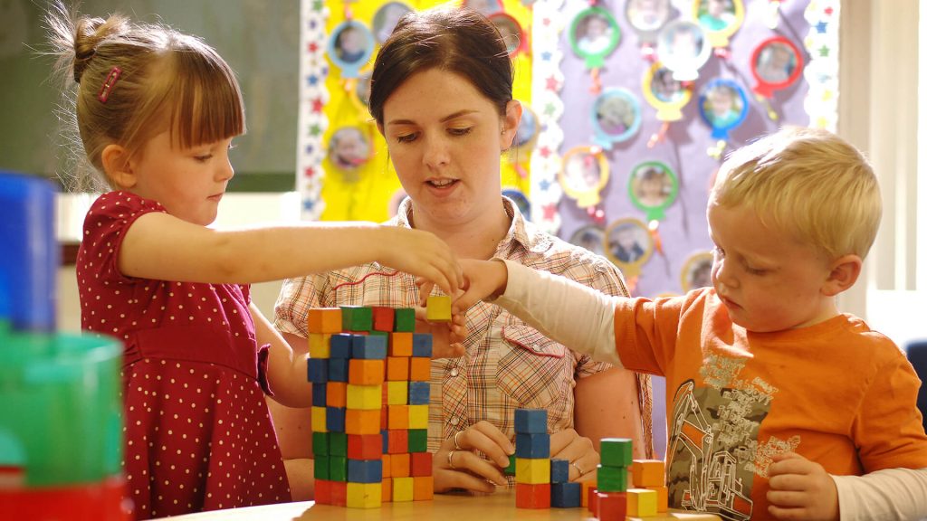 An early years professional plays with two children, stacking small buildings blocks on a table, in an early years setting.