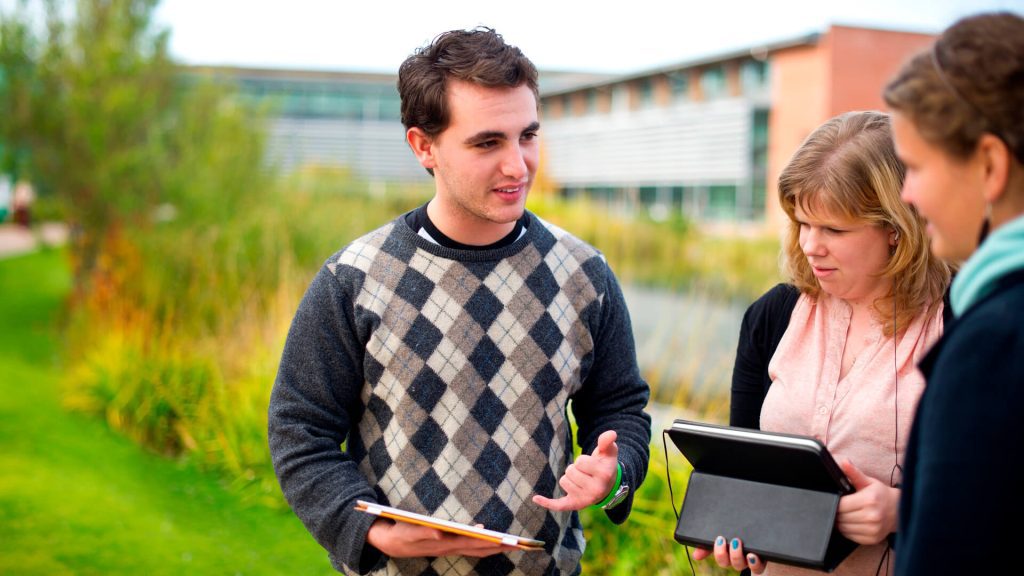 Three students have a discussion by a lake with the Faculty of Education building in the background.