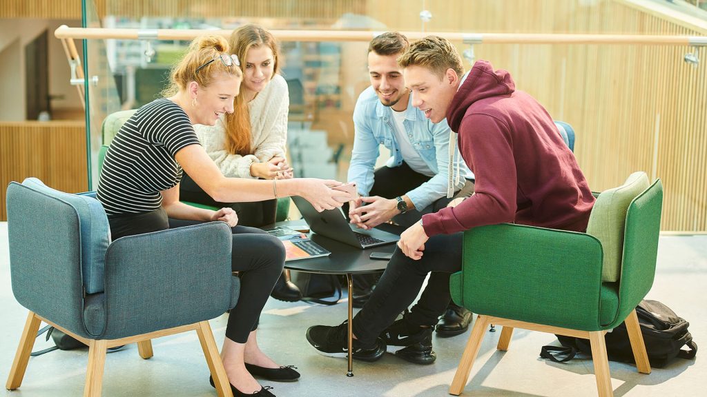 Four students sitting round a small table in the Catalyst building, with one student showing something on their mobile phone to the other three.