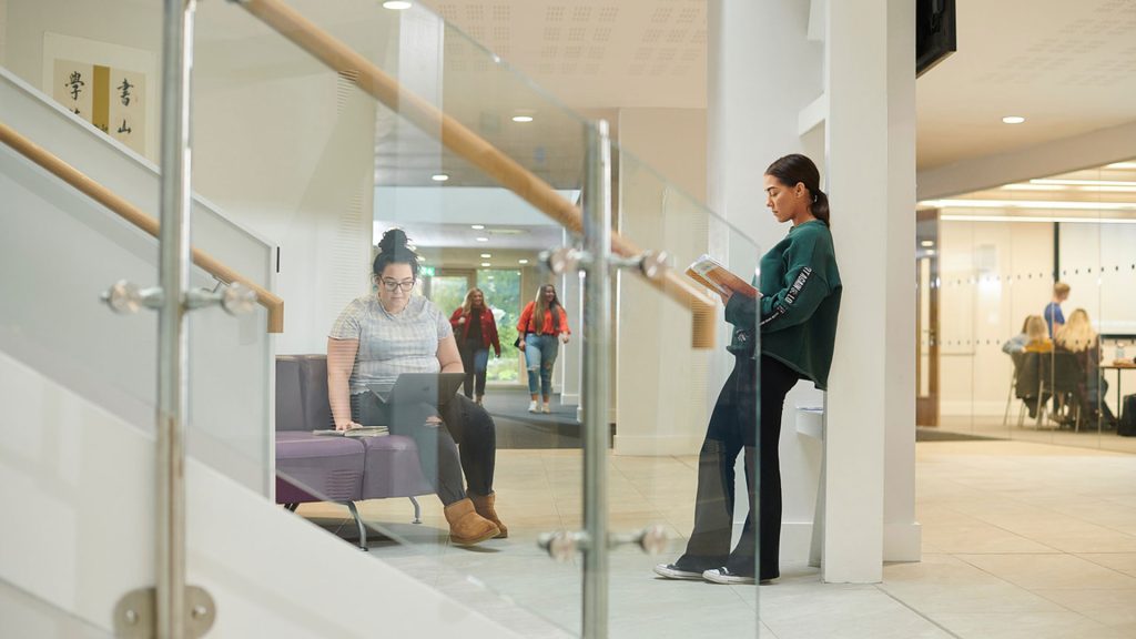 A student leans on a pillar whilst reading a textbook in the Law & Psychology building. Another student is sat on a purple leather couch with their laptop on their lap.