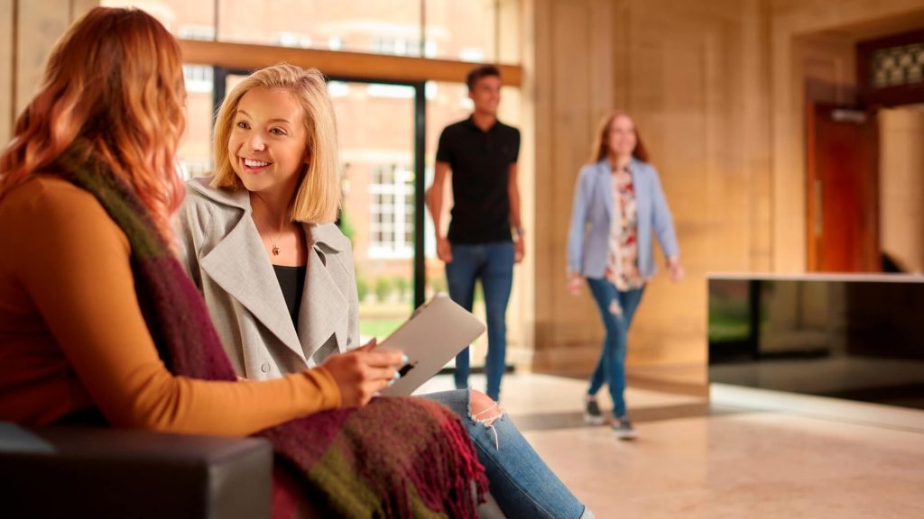 Two students chat while sitting in the reception of the Main Building. Two students walk through the entrance doors behind them.