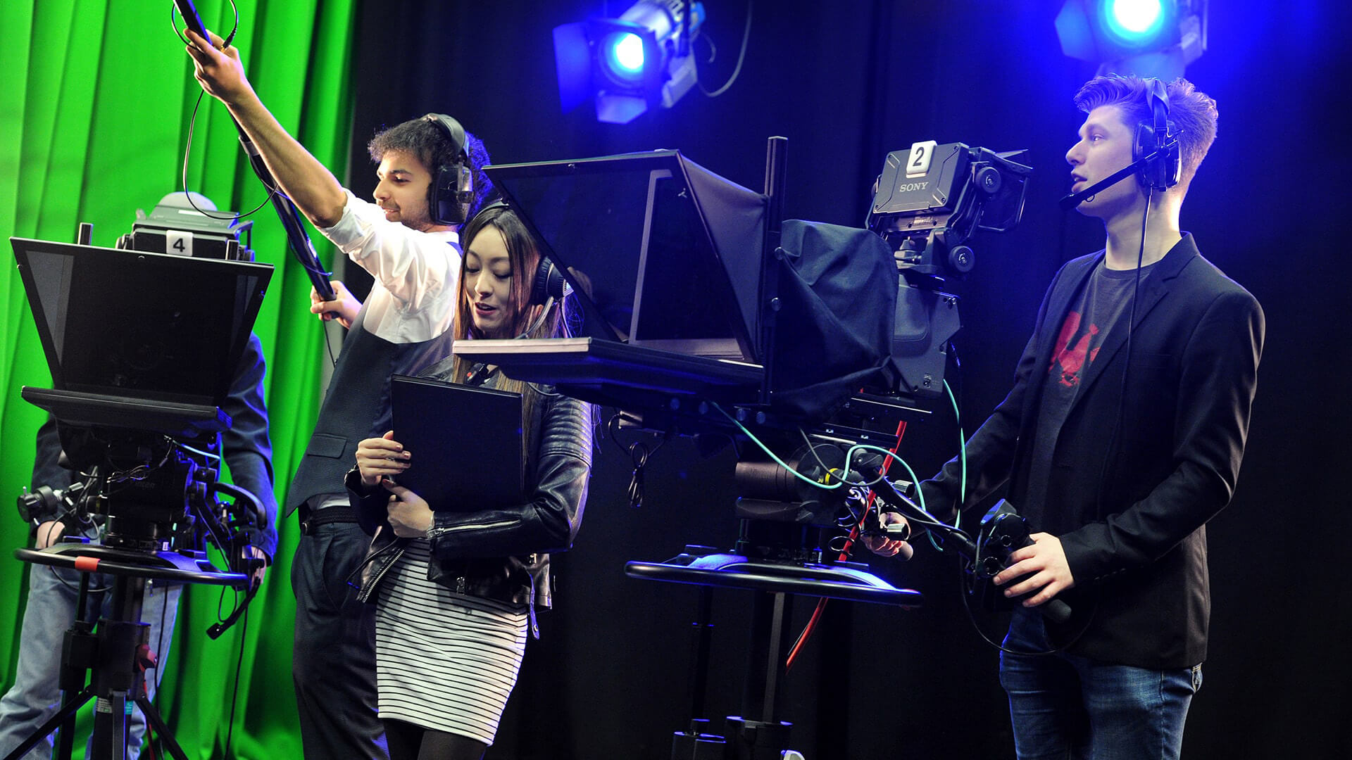 Four students film in the broadcast studio, some operating equipment and another holding a folder and supervising the exercise.