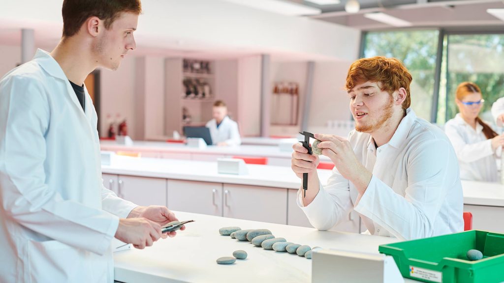 A student holds a clast and measures the size using callipers while another student observes. A tray of clasts requiring measurement is on the benchtop. Other students are working in the background doing various lab activities.