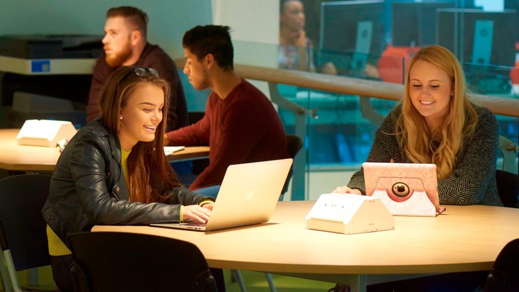 Students sit round desks working in a social learning space in the Tech Hub.