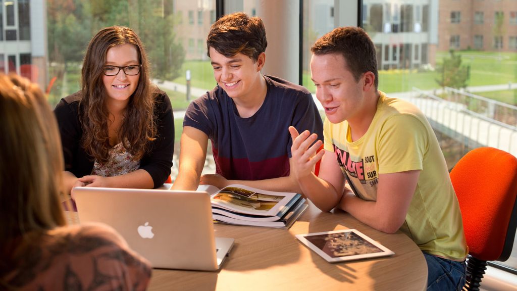 Four students sit round a table in a classroom in Creative Edge, studying a Macbook screen with textbooks open in front of them.