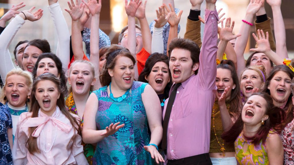 Musical theatre students, many with arms aloft, perform a song.