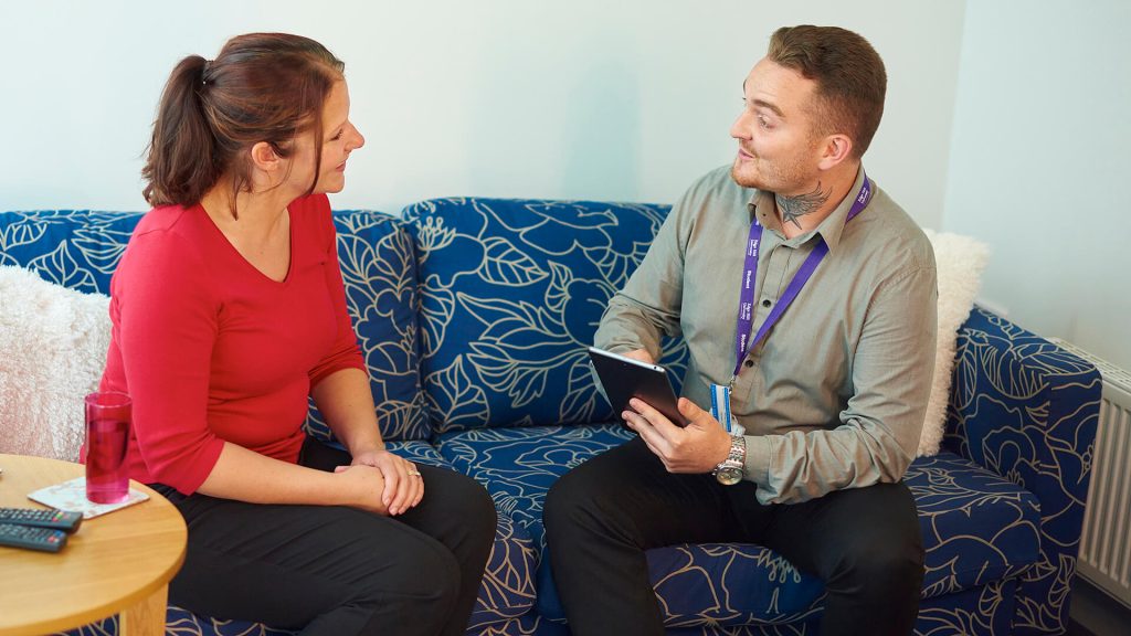 A student nurse sits on a settee and talks to a patient in a simulated home environment.