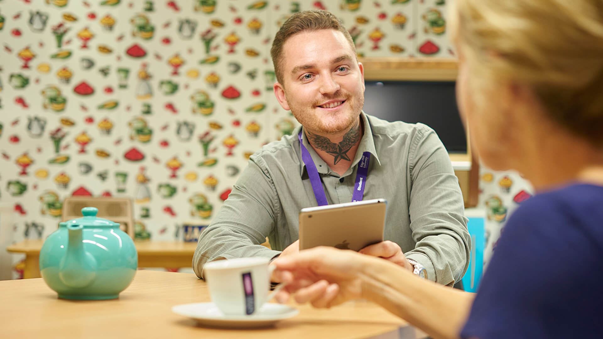 A student nurse sits at a table with a patient, who is drinking a cup of tea, and talks with them in a simulated home environment.
