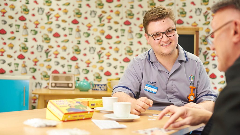 A student nurse talks with an elderly patient in a simulated home environment.