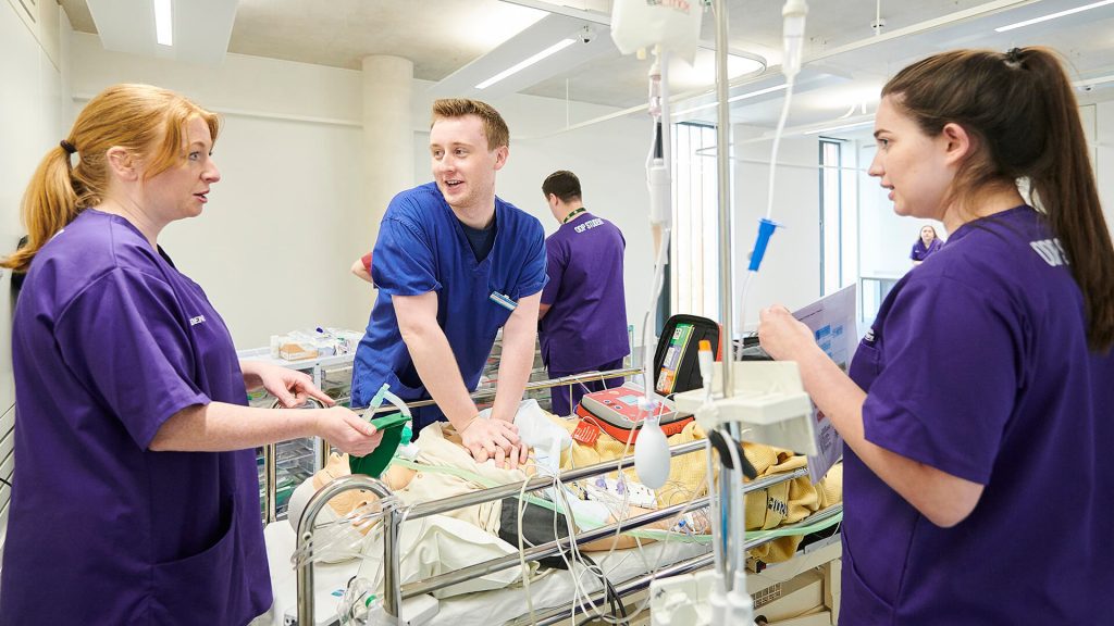 A student operating department practitioner administers CPR to a patient on a trolley, while two of his peers observe.