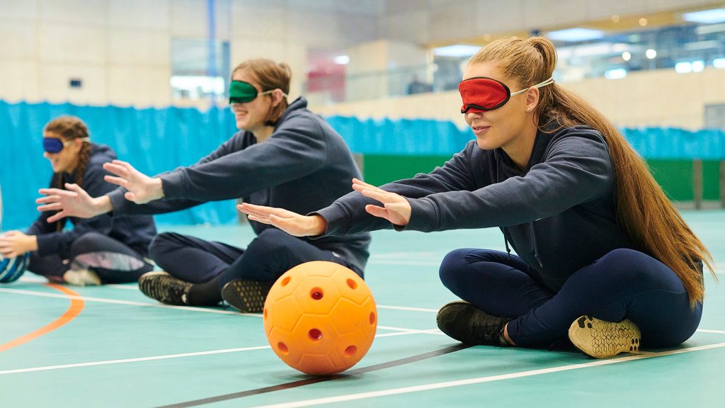 Physical Education students sit cross-legged and blindfolded on the floor, reaching out to touch a ball.