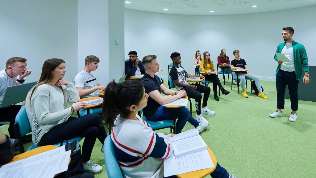 A lecturer addresses a group of students in a classroom.