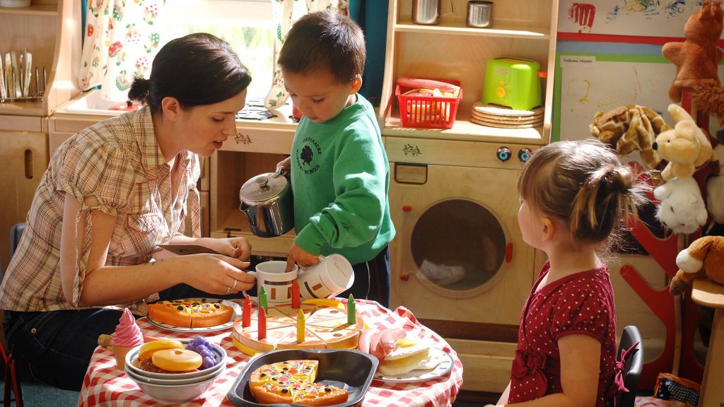 A teacher and two young children serve pretend pizza, cake and other food in an early years setting.