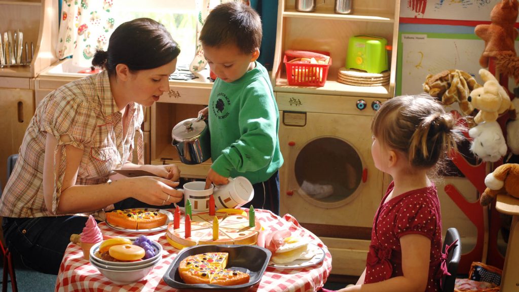 A teacher and two young children serve pretend pizza, cake and other food in an early years setting.