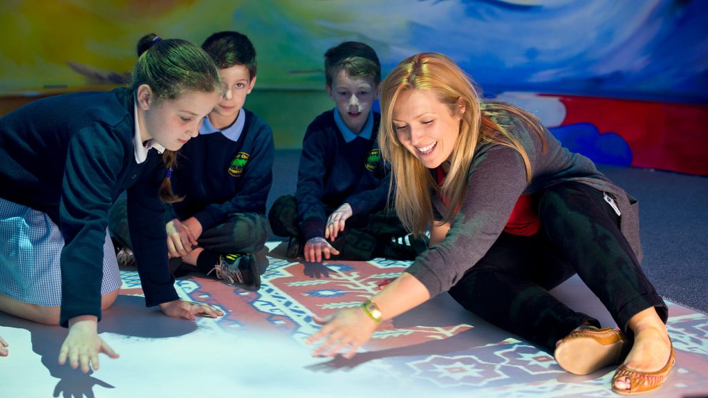 A teacher shows primary children a projection on a classroom carpet.