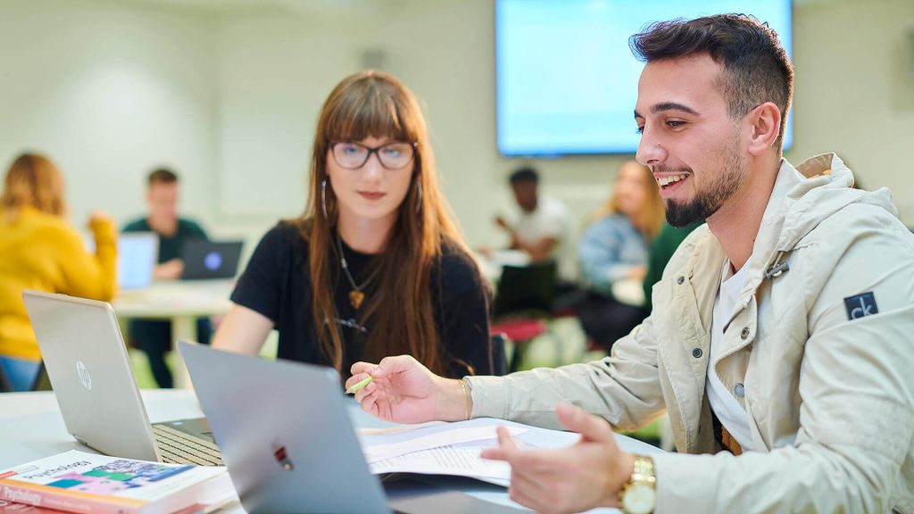 Two students chat while working on laptops in a seminar.