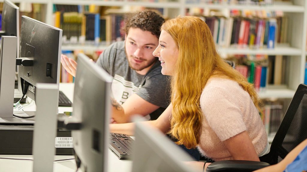 Two students talk while using computers in the University library.