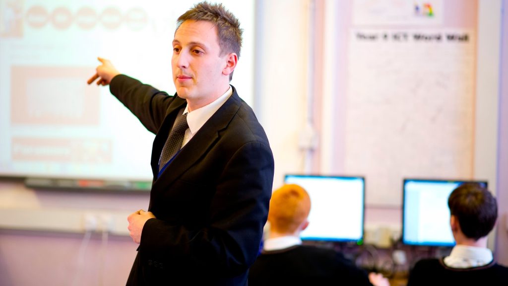 A secondary information technology teacher addresses pupils in a computing classroom.