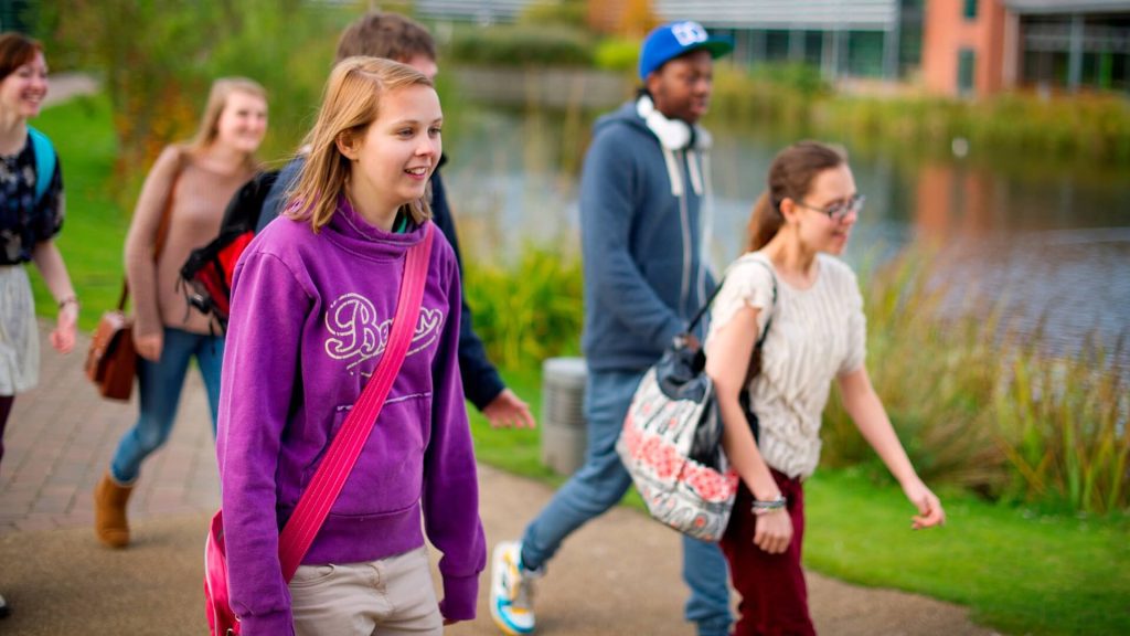 Six students walk past a lake at the western side of campus, with the Faculty of Education building in the background.