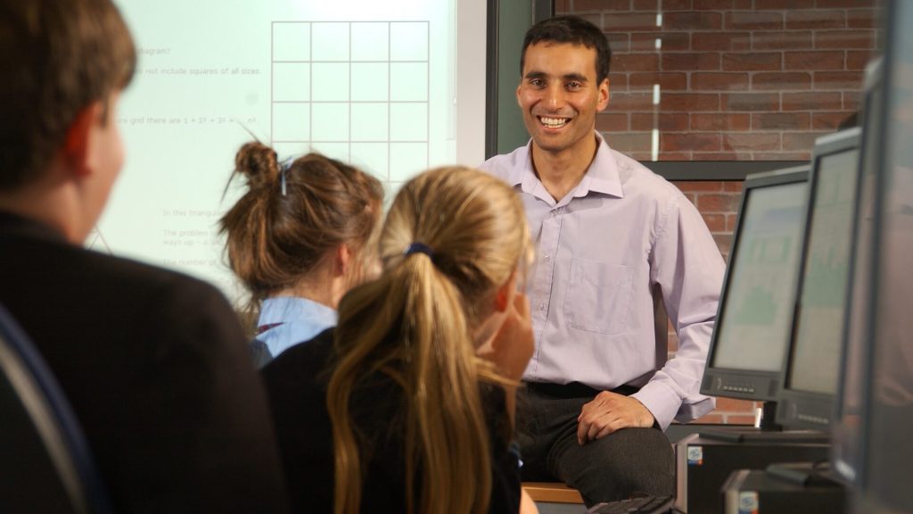 A secondary teacher addresses three pupils in a classroom who are sat in front of computers.