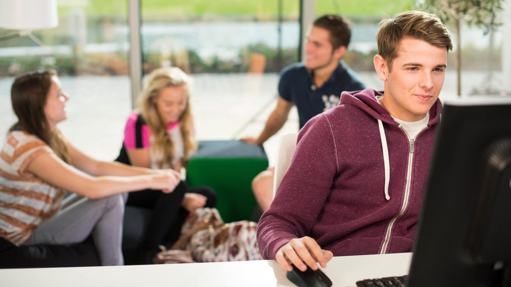 A student works at a computer in Creative Edge, with other students sat chatting behind them.