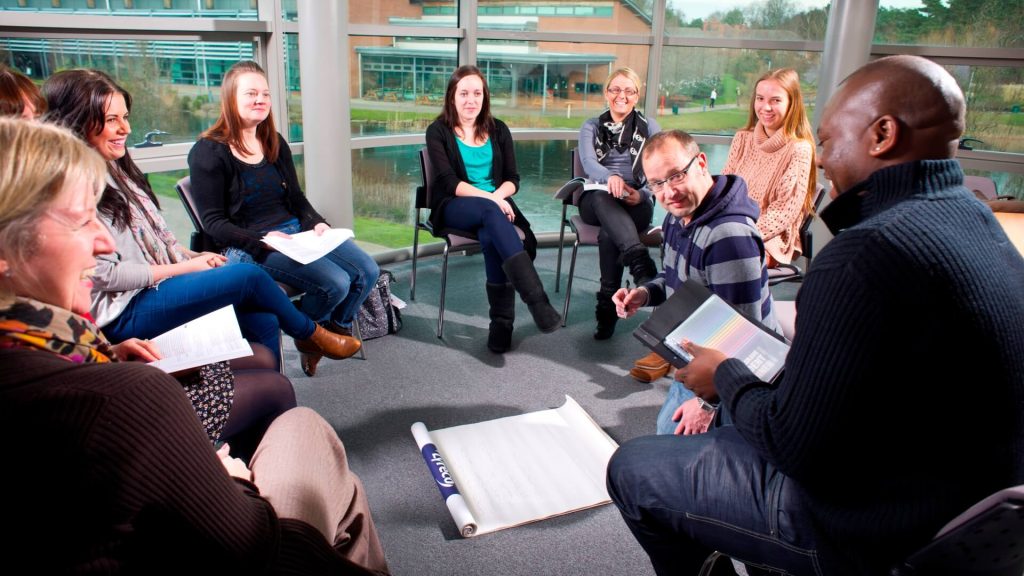 Students sit in a circle around a lecturer who is kneeling on the floor making notes on a flip chart.