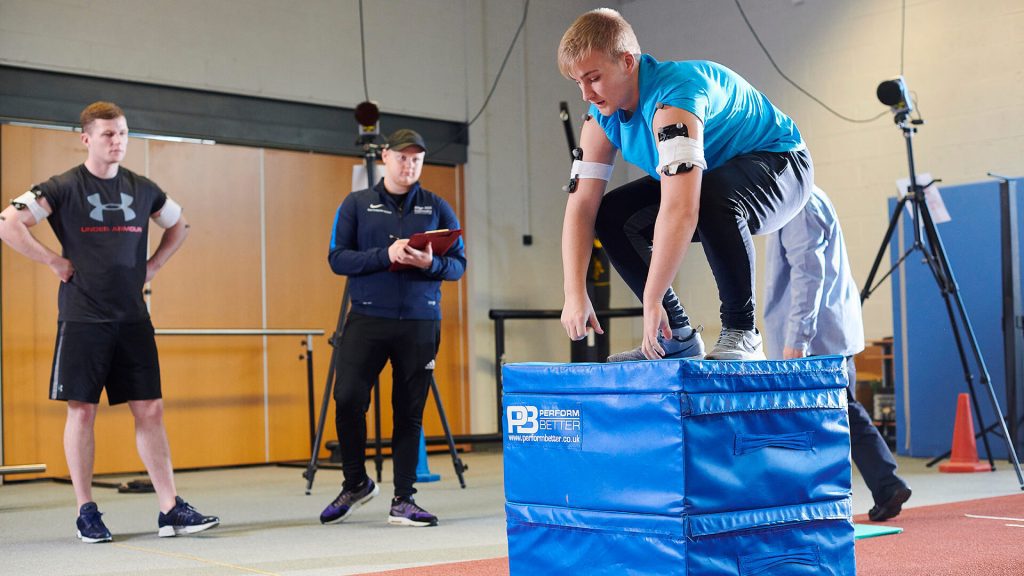 A student lifts weight while wearing monitoring equipment in a sports laboratory.