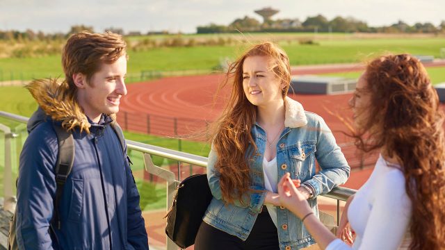 Three students chat while standing on the viewing platform on the roof of the Sports Centre which overlooks the athletics track.