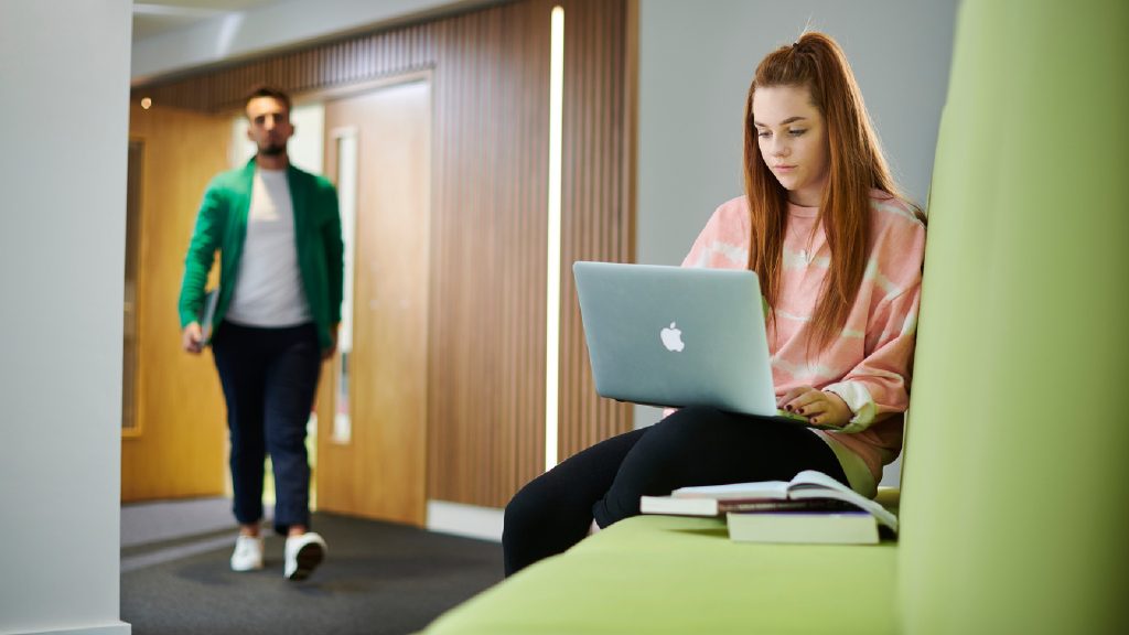 A student sat on a green couch whilst typing on their laptop. They have their textbooks next to them on the couch. There is another student walking past in the background.