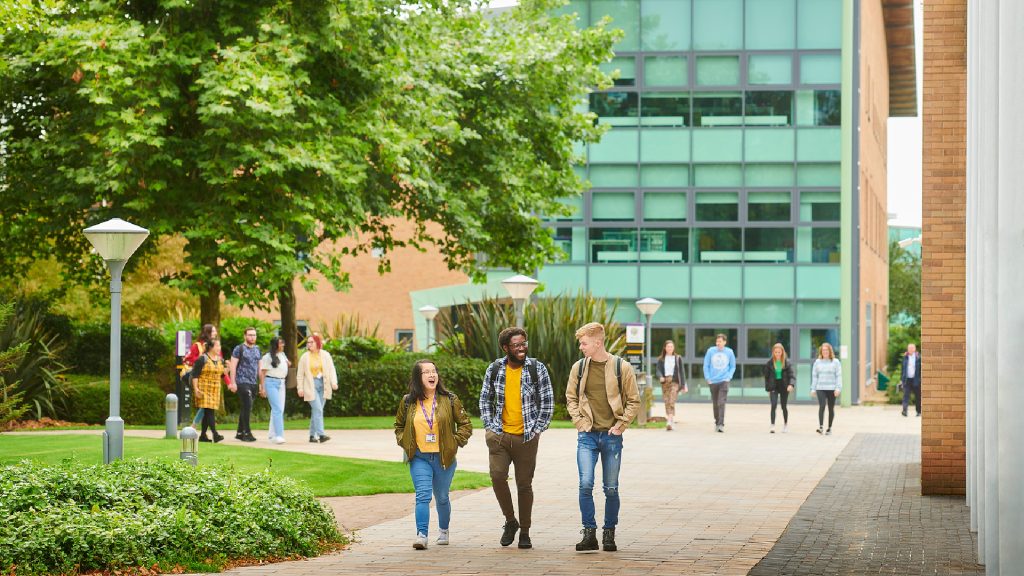 3 students walking through campus with the Clinical Skills and Simulation Centre. There are other students together in the background.