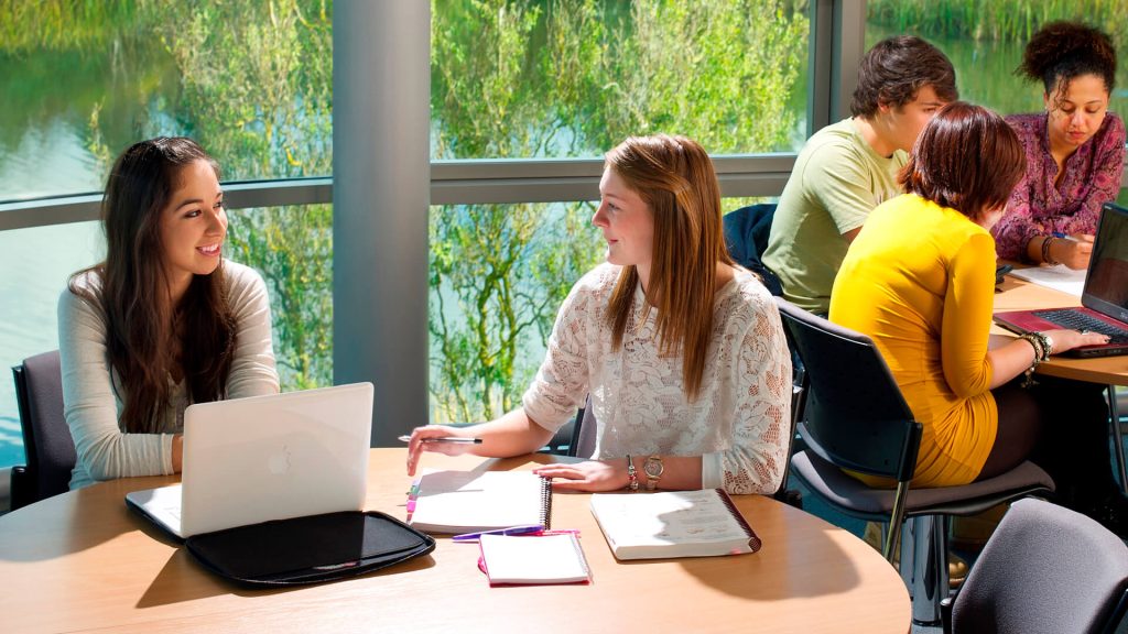 Students sitting round a desk participate in a seminar with a lake in the background.