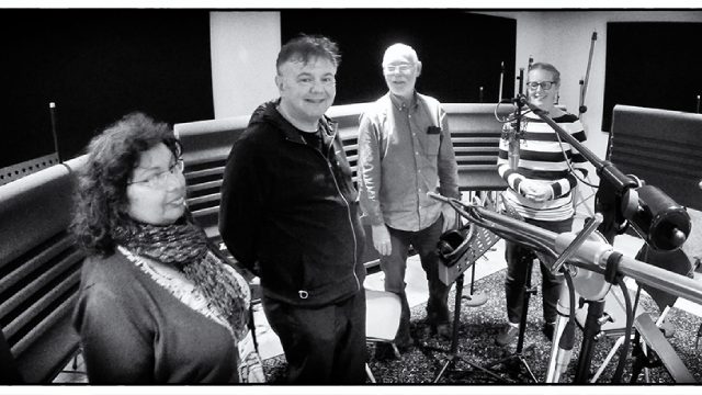A black and white image of four people in a recording studio. There are various music instruments and pieces of equipment in front of them.
