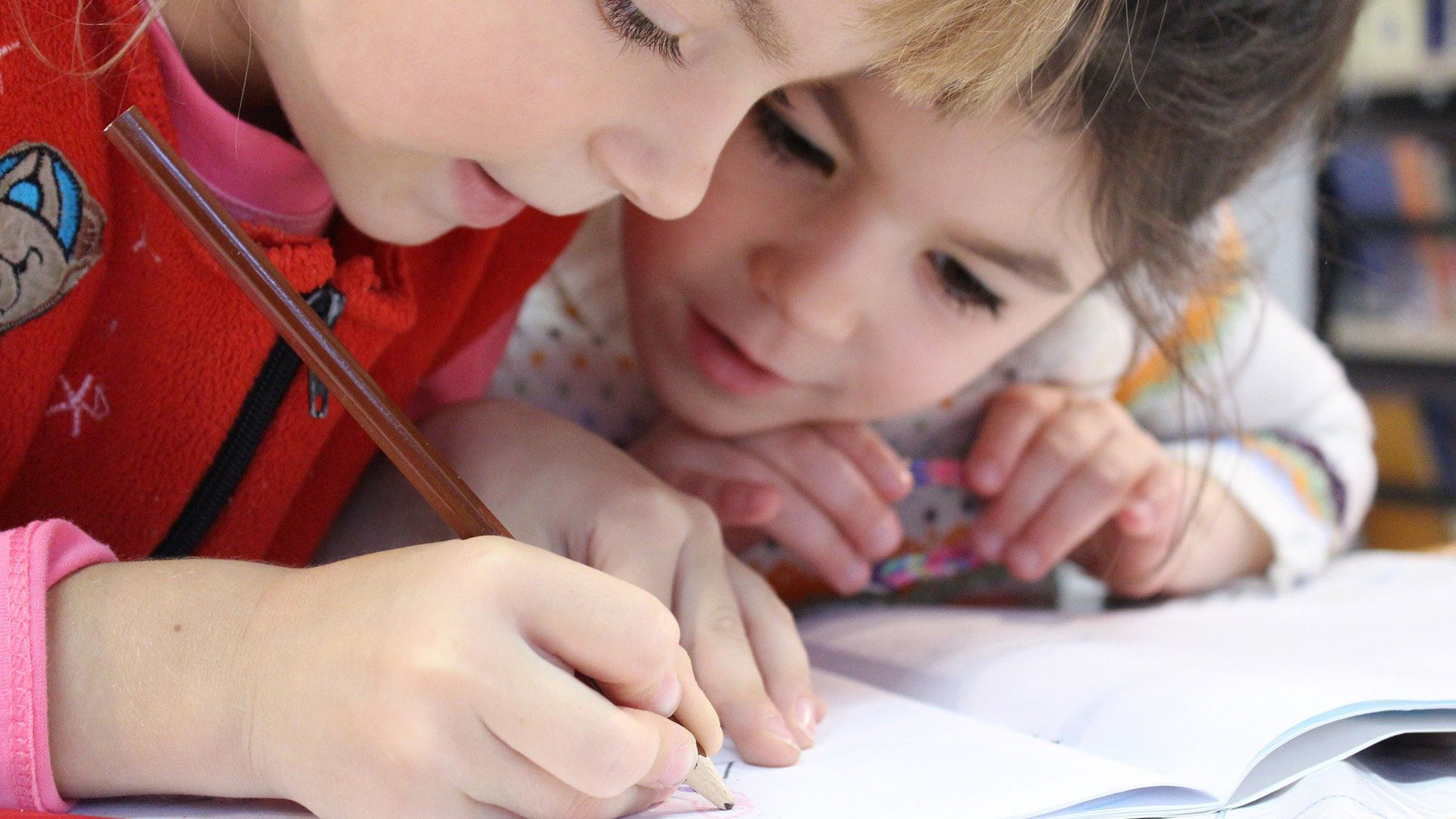Young children looking closely at a piece of paper, one child holds a pencil and is writing on the paper