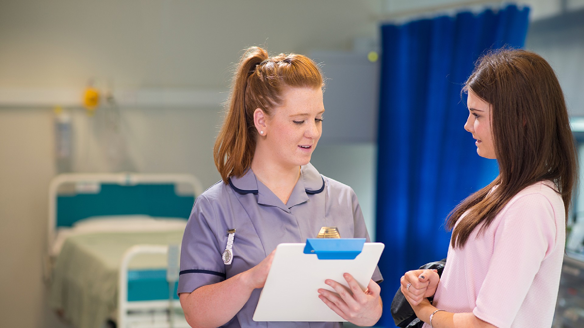 A student nurse is talking to a patient. The nurse is reading from a clipboard. There is an empty hospital bed in the background and a blue curtain to the right of it.