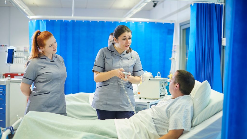 Two student nurses are stood over a hospital bed talking to a patient. One of the nurses is holding a device that is connected to the bed.