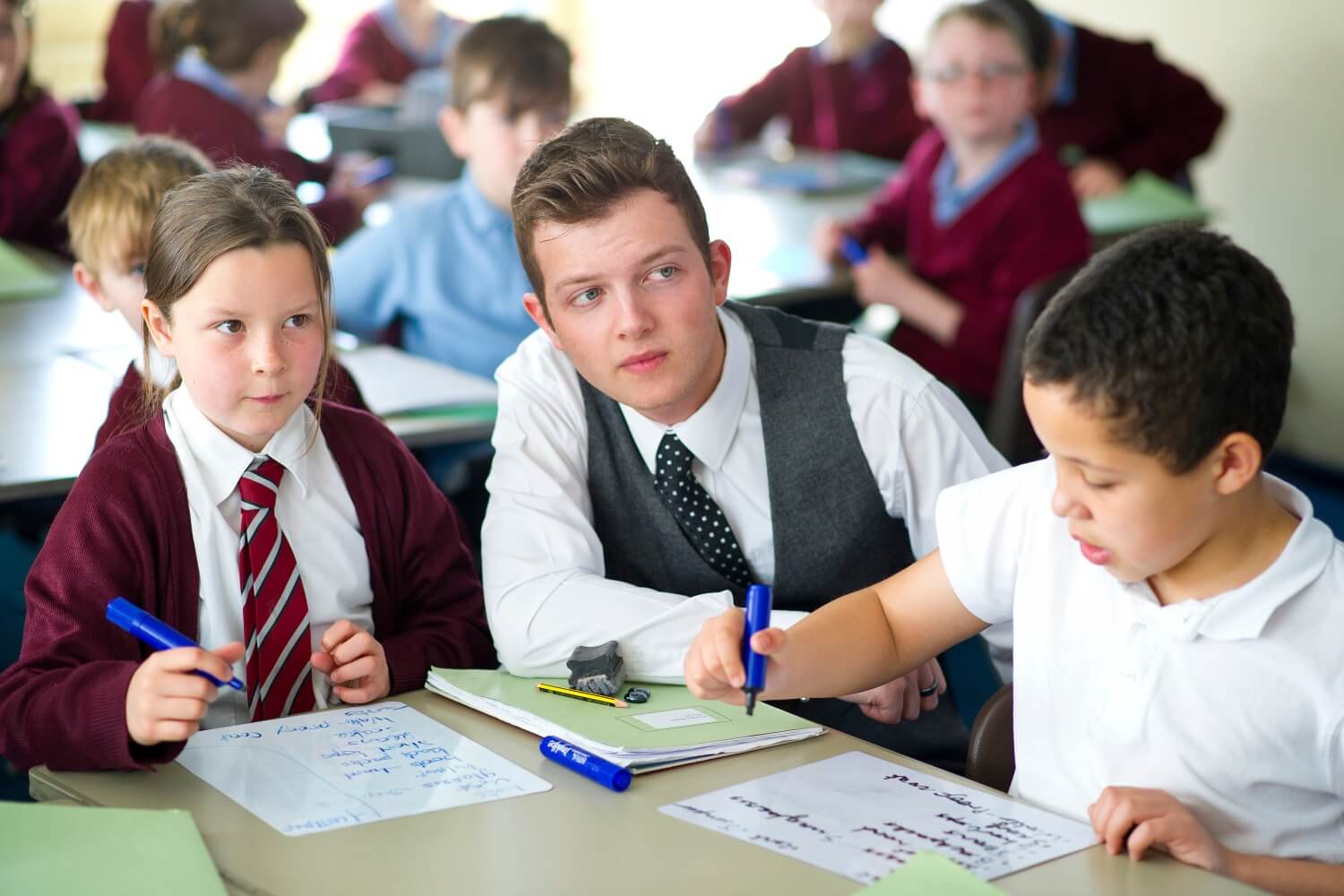 A teacher crouches down by the desk of two pupils who are writing on paper in a primary school classroom.