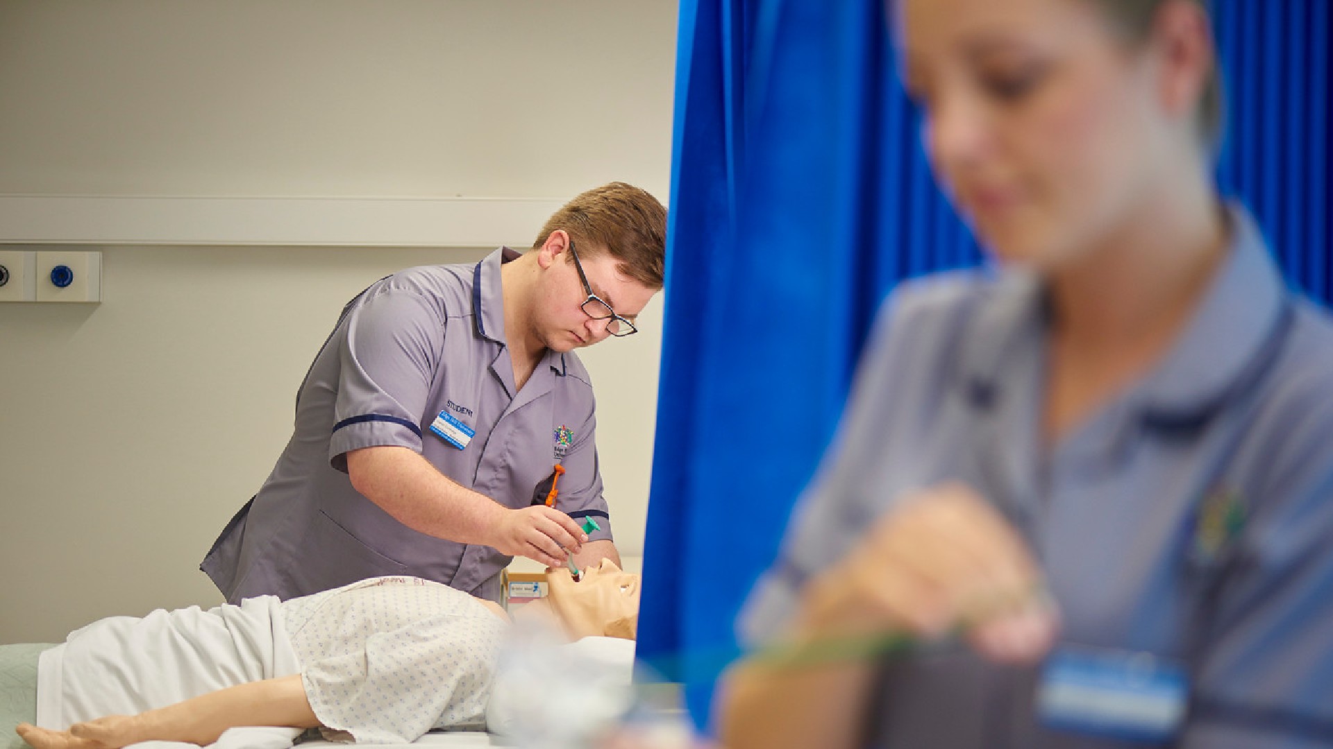 A student nurse is inserting a green tube into the mouth of a practice mannequin in the background of this image. In the foreground there is another student nurse.