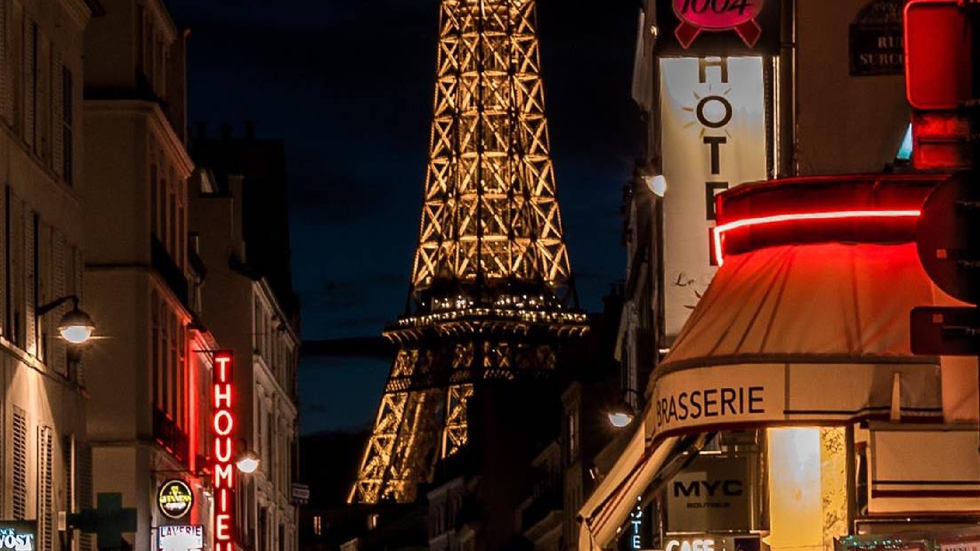 An image of a street in Paris, France. It is night and there are various lit building signs. In the background of the image the eiffel tower is visible, it is well lit.