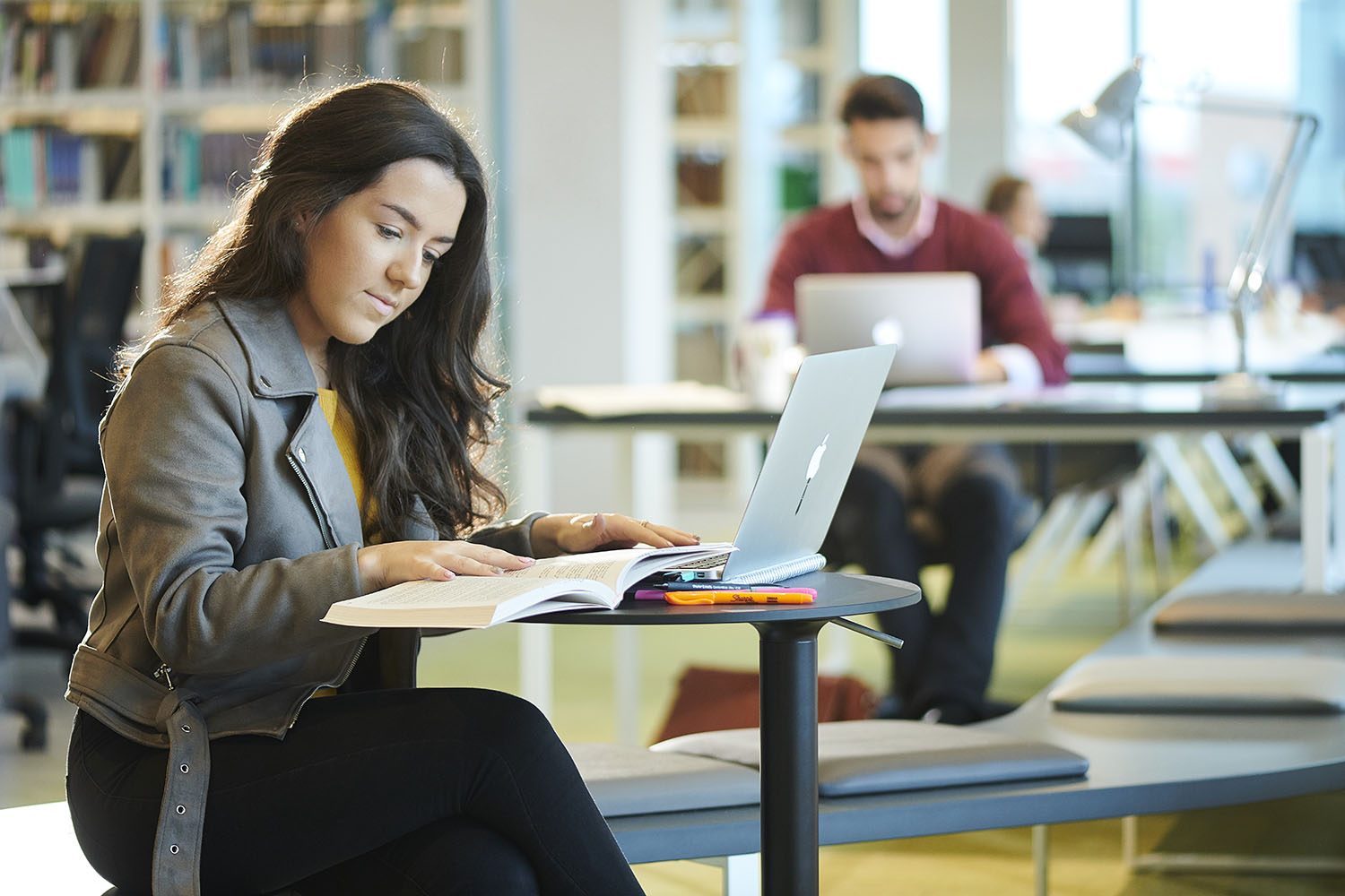 A student refers to a book while working on her Macbook in the University library.