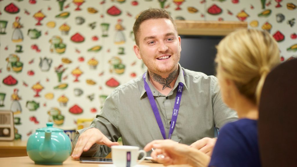 Two people are talking whilst sat at a table. There is a coloured wall behind them with various food and drinks on. There is a blue teapot on the table in front of the people. They are both looking at each other, smiling.