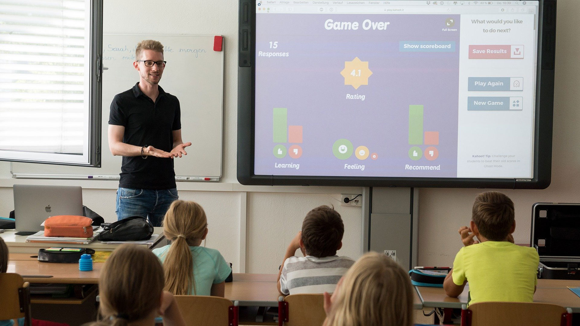 Teacher at the front of the classroom in front of an interactive board with a game on it, children are looking at the screen