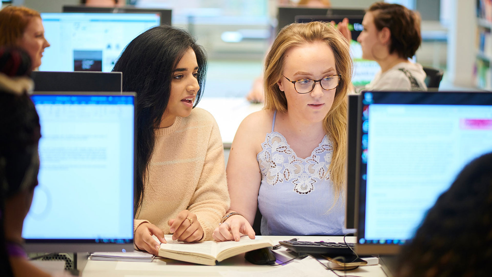 Two students in a computer lab looking at a book and a computer